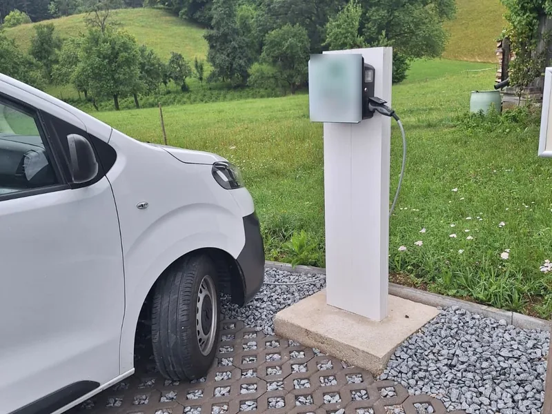 Ladesäule für Elektroautos neben einem geparkten Transporter, dahinter Wiese und bewaldete Hügel